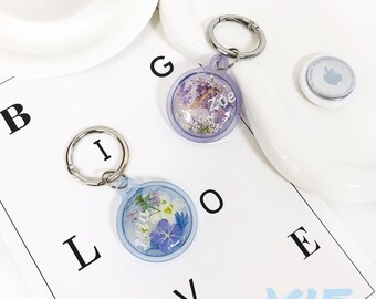 May include: Two keychains with clear round charms filled with pressed flowers and a silver ring. The charms have the names "Zoe" and "Ava" written on them. The keychains are on a white surface with the letters "B I G L O V E" written on it.