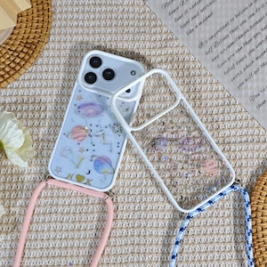 May include: Two phone cases with space-themed designs. One case is clear with glitter and the other has a light blue background with planets, stars, and the letters H and K. Both cases have a white frame and a lanyard. One lanyard is pink and the other is blue.