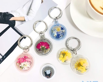 May include: A collection of clear, circular keychains with dried flowers encased inside. The keychains feature silver rings and a variety of flower colors, including pink, yellow, and blue. An Apple AirTag is also visible.