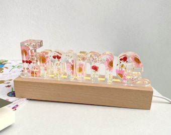 May include: A wooden base with a clear acrylic light-up sign that spells "Emma" with pressed flowers inside. The flowers are pink, red, and white.