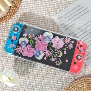May include: A clear case for a gaming console with a black background, adorned with pressed pink and purple flowers. The console has blue and red controllers. The word "Kay" is visible on the case.