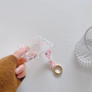 May include: Clear phone case with a floral design featuring pink blossoms and green stems. The case has a beaded wrist strap with pink beads and a gold-colored ring. The case is held in a hand.