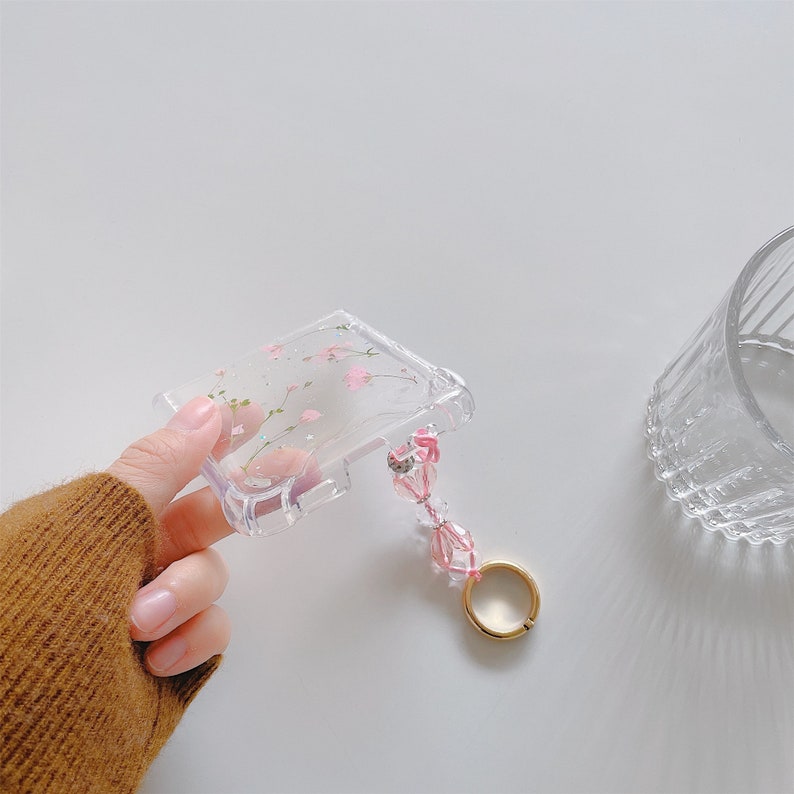 May include: Clear phone case with a floral design featuring pink blossoms and green stems. The case has a beaded wrist strap with pink beads and a gold-colored ring. The case is held in a hand.
