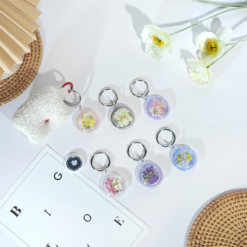 May include: A collection of clear, round keychains filled with dried flowers in various colors. Each keychain has a silver-colored ring. A white fluffy pouch with a red loop is also present, along with a white paper with letters.