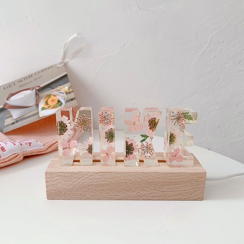 May include: A wooden base with a light wood finish holds five clear resin letters spelling out the name "MIKE". Each letter is filled with pressed pink and green flowers.