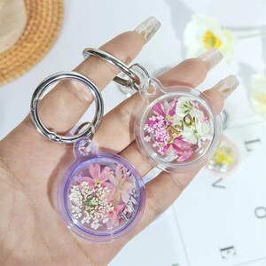 May include: Two clear resin keychains with dried flowers and silver hardware. One keychain has a purple border and the word "Love" in silver. The other keychain has the name "Ava" in gold. Both keychains have a round shape.