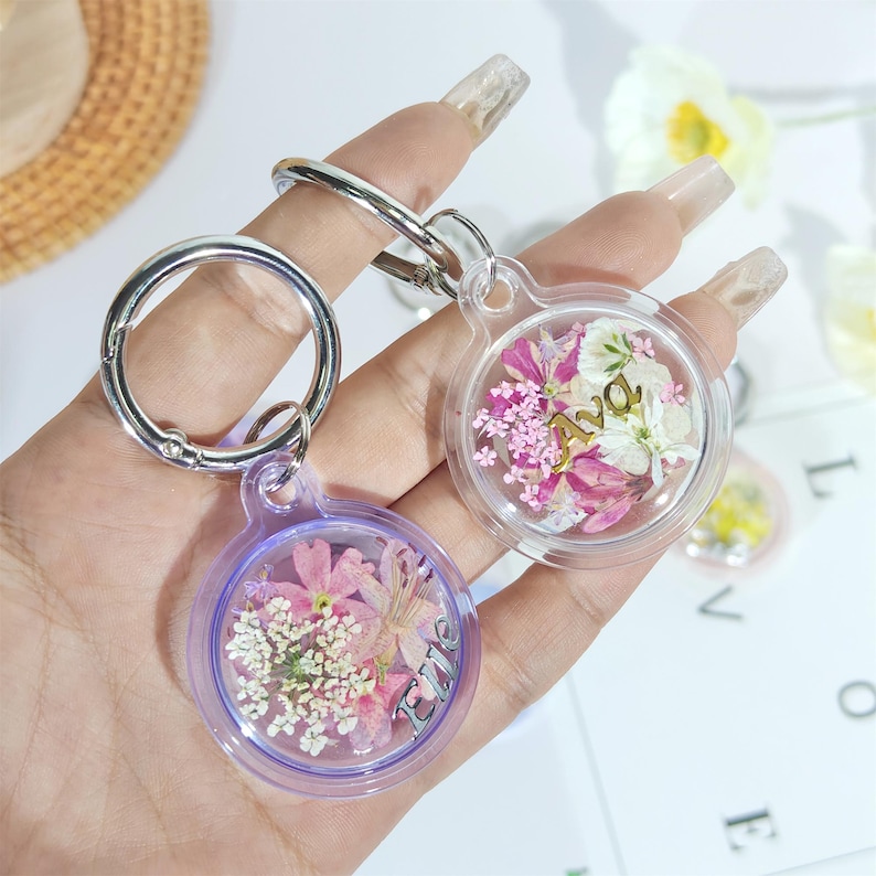 May include: Two clear resin keychains with dried flowers and silver hardware. One keychain has a purple border and the word "Love" in silver. The other keychain has the name "Ava" in gold. Both keychains have a round shape.
