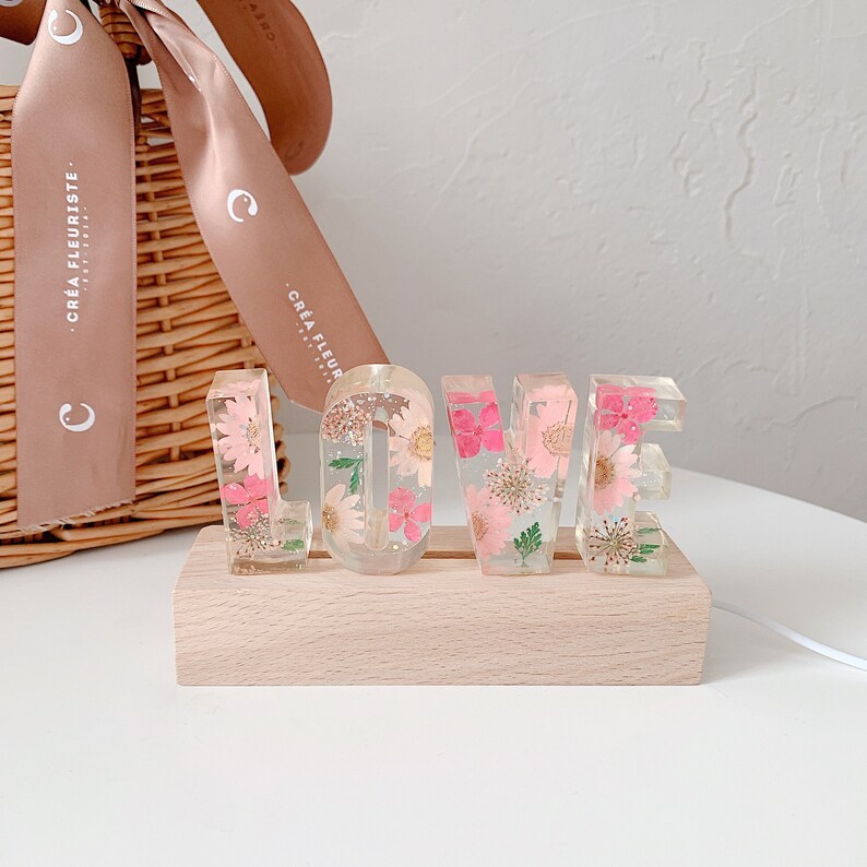 May include: A wooden base with a clear resin "LOVE" sign. The letters are filled with pressed pink and white flowers. The sign is lit up with warm white lights.