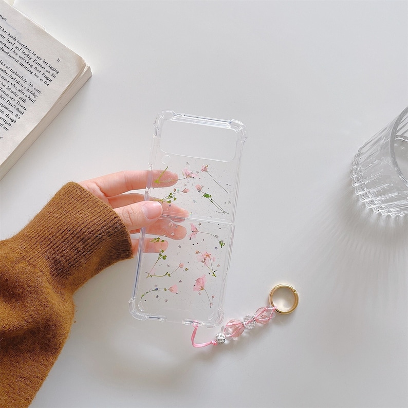 May include: Clear phone case with a floral design featuring pink flowers and silver glitter. The case has a pink lanyard with a beaded charm and a gold ring. The phone case is open, revealing the phone's interior.