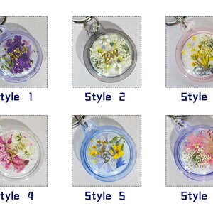 May include: A collection of six clear resin keychains, each with a different arrangement of dried flowers and decorative elements. Each keychain is labeled with a style number. The keychains have silver-colored key rings.