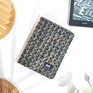 May include: A textured, multi-colored book cover with a small blue label that reads "MOON". The cover is made of a woven fabric with shades of blue, beige, and black. A Kindle e-reader is visible in the background.