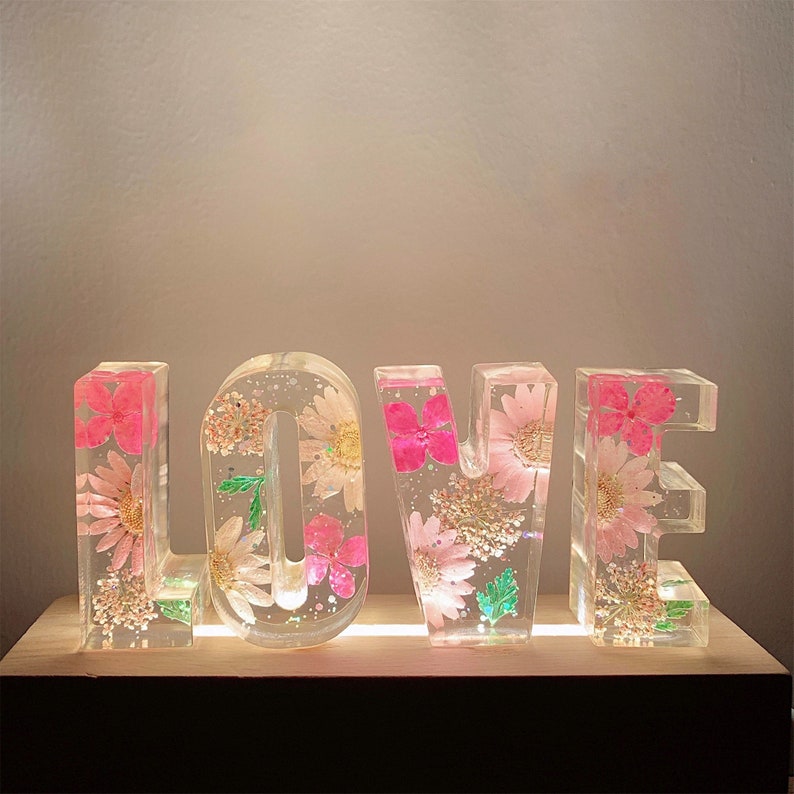 May include: A decorative light-up sign spelling "LOVE" with pink and green pressed flowers encased in clear resin.