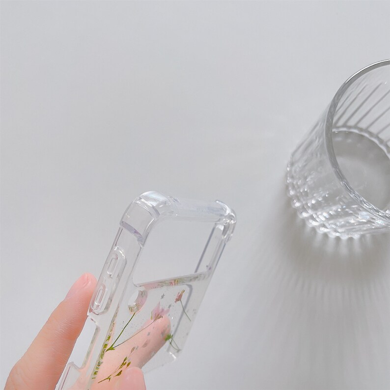 May include: Clear phone case with a floral design. The case is transparent, allowing the colorful flowers and green stems to be visible. The case is held in a person's hand, and a clear glass is in the background.