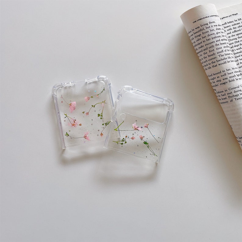 May include: Two clear phone cases with a floral design. Each case features small pink flowers and green sprigs encased within the transparent plastic. Silver glitter adds a touch of sparkle to the design.