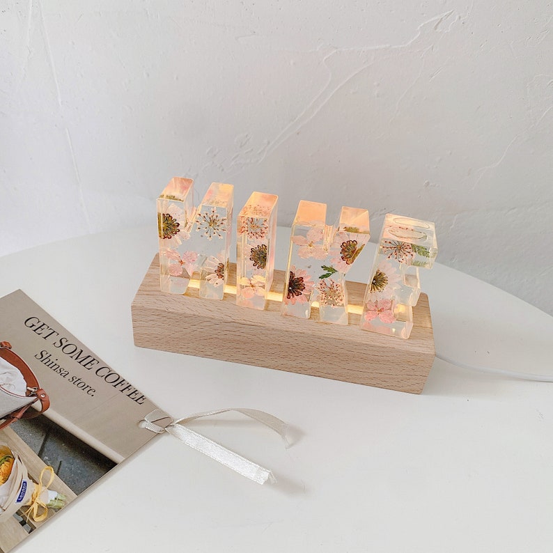 May include: A wooden base with a light switch holds five clear resin letters spelling "MIKE" with dried flowers pressed inside. The letters are illuminated from behind, casting a warm glow.