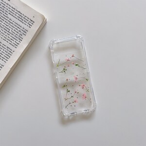 May include: Clear phone case with embedded pink flowers and silver glitter. The case is designed to fit a folding phone. The floral design adds a touch of nature and elegance to the phone's appearance.