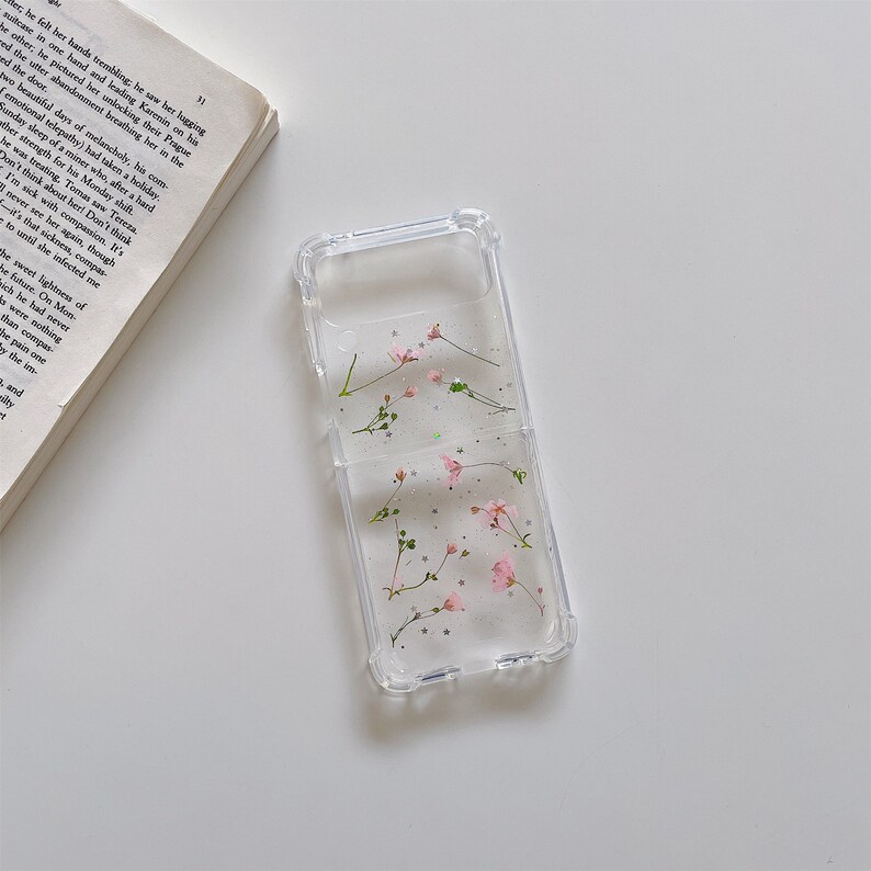 May include: Clear phone case with embedded pink flowers and silver glitter. The case is designed to fit a folding phone. The floral design adds a touch of nature and elegance to the phone's appearance.