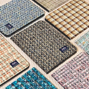 Puede incluir: Una variedad de fundas cubiertas de tela en varios colores y patrones. Las fundas presentan un material texturizado similar al tweed en tonos de azul, marrón, rosa y beige. Cada funda tiene un borde negro y una pequeña etiqueta con un logotipo.
