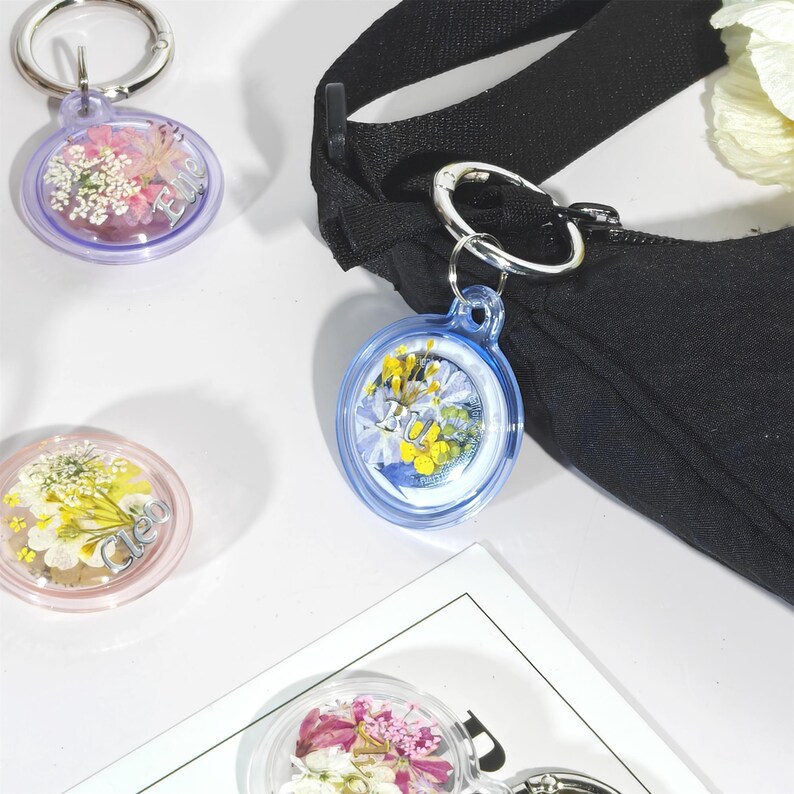 May include: Clear resin keychains with dried flowers and silver-colored key rings. The keychains are round and feature a variety of colorful dried flowers. One keychain has the letters "RU" in yellow. Another keychain has the name "Cleo".