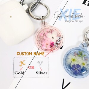 Puede incluir: Un llavero de resina transparente y rosa con un peque&ntilde;o ramo de flores secas en su interior. El llavero tiene un dije plateado con la letra "X" en &eacute;l. El texto "CUSTOM NAME" es visible debajo del llavero. Las palabras "Gold" y "Silver" son visibles debajo del dije. La palabra "OR" es visible entre las palabras "Gold" y "Silver".