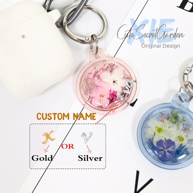 Puede incluir: Un llavero de resina transparente y rosa con un peque&ntilde;o ramo de flores secas en su interior. El llavero tiene un dije plateado con la letra "X" en &eacute;l. El texto "CUSTOM NAME" es visible debajo del llavero. Las palabras "Gold" y "Silver" son visibles debajo del dije. La palabra "OR" es visible entre las palabras "Gold" y "Silver".