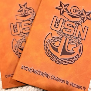 May include: Two tan leather-like notebooks featuring a dark brown embossed USN anchor emblem with stars and chains. The front cover displays the text AVCM(AW/SW/IW) Christian W. Hansen IV, while the second notebook shows the text CMC.