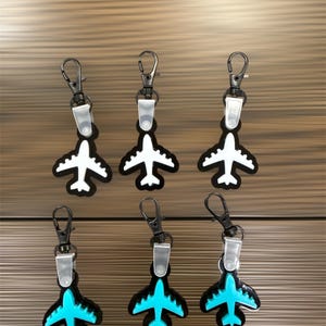 May include: Six airplane-shaped keychains. Three have white planes on a black background, and three have turquoise planes on a black background. Each keychain has a silver clasp and a clear plastic clip.
