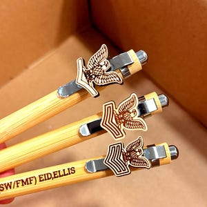 May include: Three bamboo pens with silver accents and wooden eagle emblems. The pens have a natural wood grain appearance and are arranged diagonally. The text "(SW/FMF) EID, ELLIS" is visible on one pen.