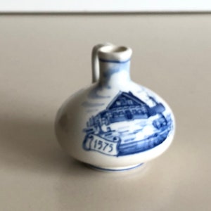 May include: A small, white ceramic jug with a blue and white design. The design features a house with a date of 1973.