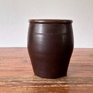 May include: A dark brown, antique-style ceramic jar with a slightly tapered shape. The jar has a rounded top and a smooth, matte finish. It sits on a wooden surface with visible wood grain.