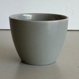 Può includere: Una tazza in ceramica di colore grigio chiaro con una forma rotonda e liscia.