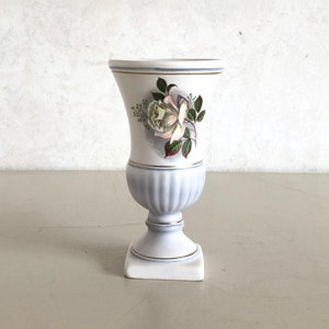 May include: A white ceramic vase with blue and gold trim and a floral design featuring a white rose. The vase has a square base and a round body.