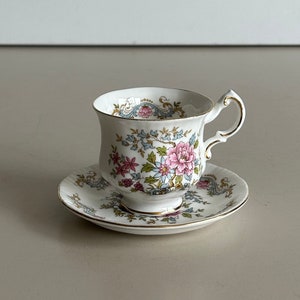 Könnte beinhalten: Ein weißes Tee-Set mit einer Tasse und Untertasse mit einem floralen Muster in Rosa, Blau und Grün. Die Tasse hat einen goldenen Henkel und Zierleisten. Die Untertasse ist rund und passt zum Design der Tasse.