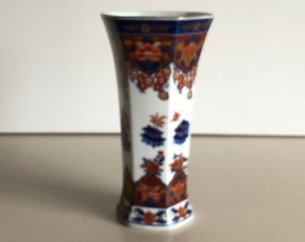 May include: A white porcelain vase with a geometric shape and a colourful floral pattern in shades of blue, red, and gold. The vase has a narrow opening and a wide base.