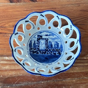 May include: A white and blue ceramic dish with a lace-like design around the edge. The center of the dish features a blue and white illustration of a castle with the text "MON. VERBALENEN 1940-1945 VOORTHUIZEN".