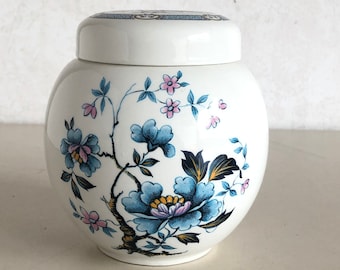 Może przedstawiać: Biały ceramiczny słoik z kwiatowym wzorem. Słoik ma niebieski i różowy wzór kwiatowy z niebieską i białą pokrywką.