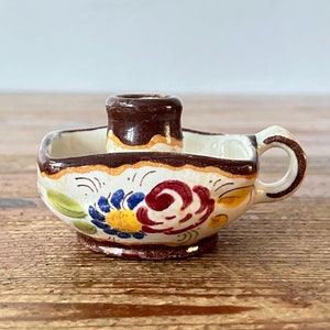 May include: A small, white ceramic candle holder with a brown rim and a floral design in blue, red, yellow, and green. The holder has a single candle opening and a handle.