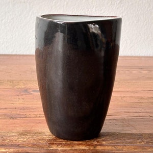 May include: A black ceramic vase with a slightly irregular, rounded shape. The vase has a glossy finish and is sitting on a wooden surface.