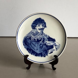 May include: A blue and white ceramic plate with a portrait of a man in a blue and white outfit. The plate is on a wooden stand.