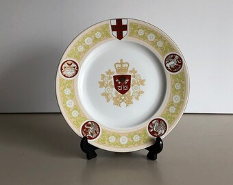 May include: A white ceramic plate with a gold and red design. The plate features a royal crest with a crown and a cross. The plate is on a black stand.