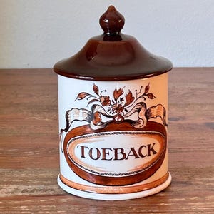 Pode incluir: Pote de cerâmica branco com tampa e base marrom. O pote apresenta um rótulo oval marrom com a palavra "TOEBACK" no centro, rodeado por elementos florais decorativos. O pote é provavelmente para guardar tabaco.
