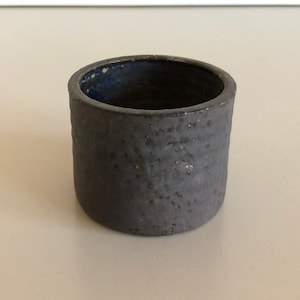 Könnte beinhalten: Ein kleiner, dunkelgrauer Keramiktopf mit einer rauen, strukturierten Oberfläche. Der Topf hat einen leicht dunkleren blauen Rand.