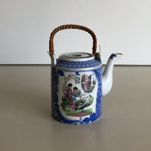 Op de afbeelding: Een klein blauw en wit porseleinen theepot met een bamboe handvat. De theepot heeft een geschilderd tafereel van twee vrouwen in traditionele kleding.
