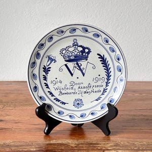 Può includere: Un piatto in ceramica bianca con design floreale e corona blu. Il piatto ha del testo ed è esposto su un supporto nero. Il design del piatto include gli anni 1914 e 1919.