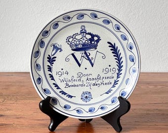 Puede incluir: Un plato de cerámica blanca con diseño floral y corona azul. El plato tiene texto y se muestra sobre un soporte negro. El diseño del plato incluye los años 1914 y 1919.