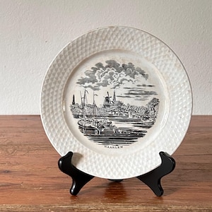 May include: White ceramic plate featuring a black and white illustration of a harbor scene with ships and buildings. The plate has a textured rim and is displayed on a black stand. The word "HAARLEM" is printed on the plate.