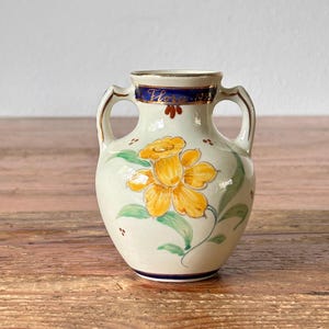 Op de afbeelding: Een kleine witte keramische vaas met twee handvatten. De vaas is versierd met een gele bloem en groene bladeren. De vaas heeft een blauwe band rond de bovenkant en de woorden "Vere 1953" in gouden letters.