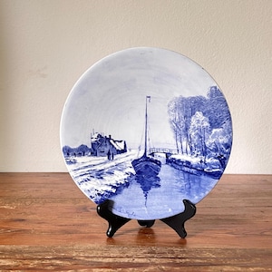 Op de afbeelding: Een blauw-wit decoratief bord met een kanaalscène met een zeilboot, brug en gebouwen. Het bord wordt gepresenteerd op een zwarte standaard tegen een houten oppervlak. Het kunstwerk is gedetailleerd en het kleurenpalet is monochroom.