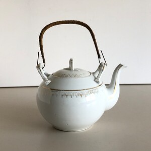 Könnte beinhalten: Weiße Porzellan-Teekanne mit einem braunen geflochtenen Henkel und goldenen Verzierungen. Die Teekanne hat einen runden Korpus und einen Ausguss an der Seite.