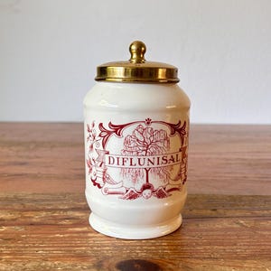 Op de afbeelding: Witte keramische pot met een gouden deksel en een rood-wit ontwerp. De pot heeft de tekst "DIFLUNISAL" erop gedrukt.